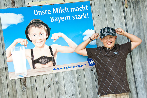 Ein Junge steht vor Plakat "Unsere Milch macht Bayern stark" und zeigt seine Muskeln.